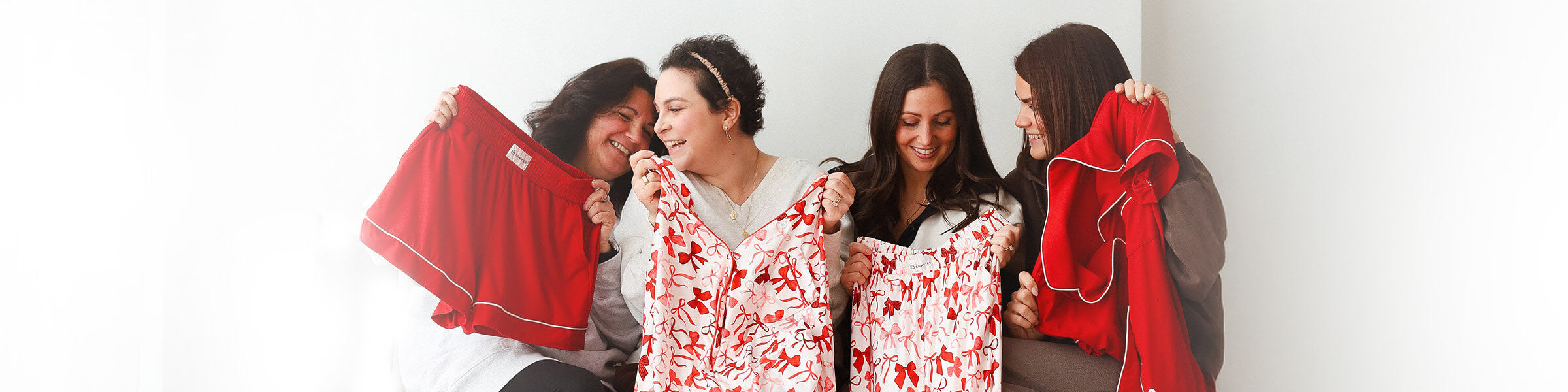 Four people sitting together and smiling while holding red and red-and-white bow-pattern pajama pieces, including shorts, pants, and a button-up top, against a bright neutral background