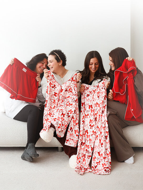 Four people sitting together and smiling while holding red and red-and-white bow-pattern pajama pieces, including shorts, pants, and a button-up top, against a bright neutral background