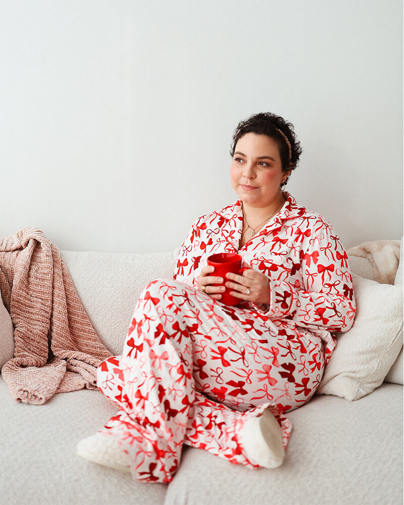 Person sitting on a couch wearing red and white bow-pattern pajamas, holding a red mug and relaxing in a cozy living room setting