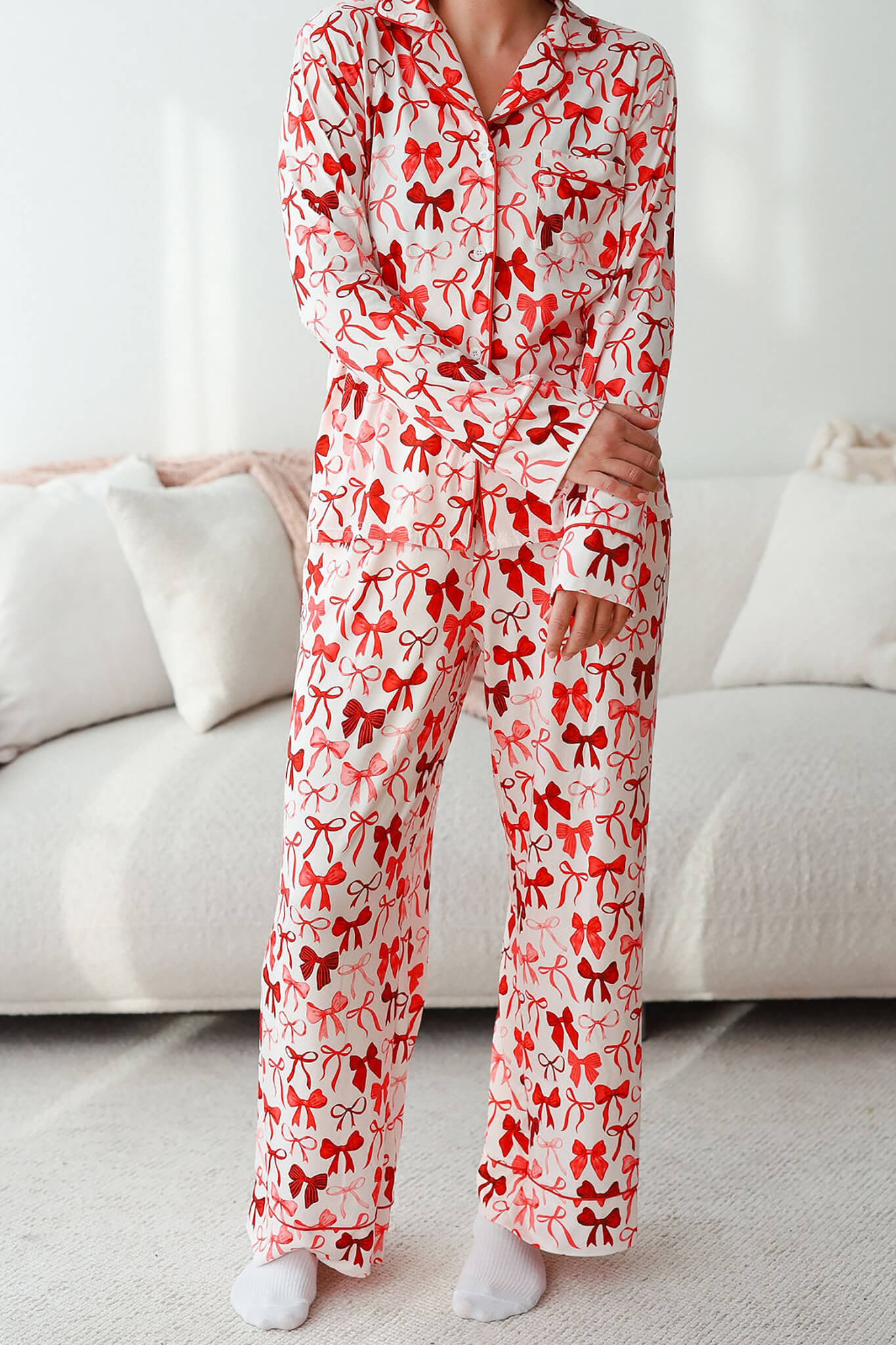Long sleeve pajama set with white fabric and red bow print, featuring a button-up top and straight-leg pants, worn with white socks in a bedroom setting.