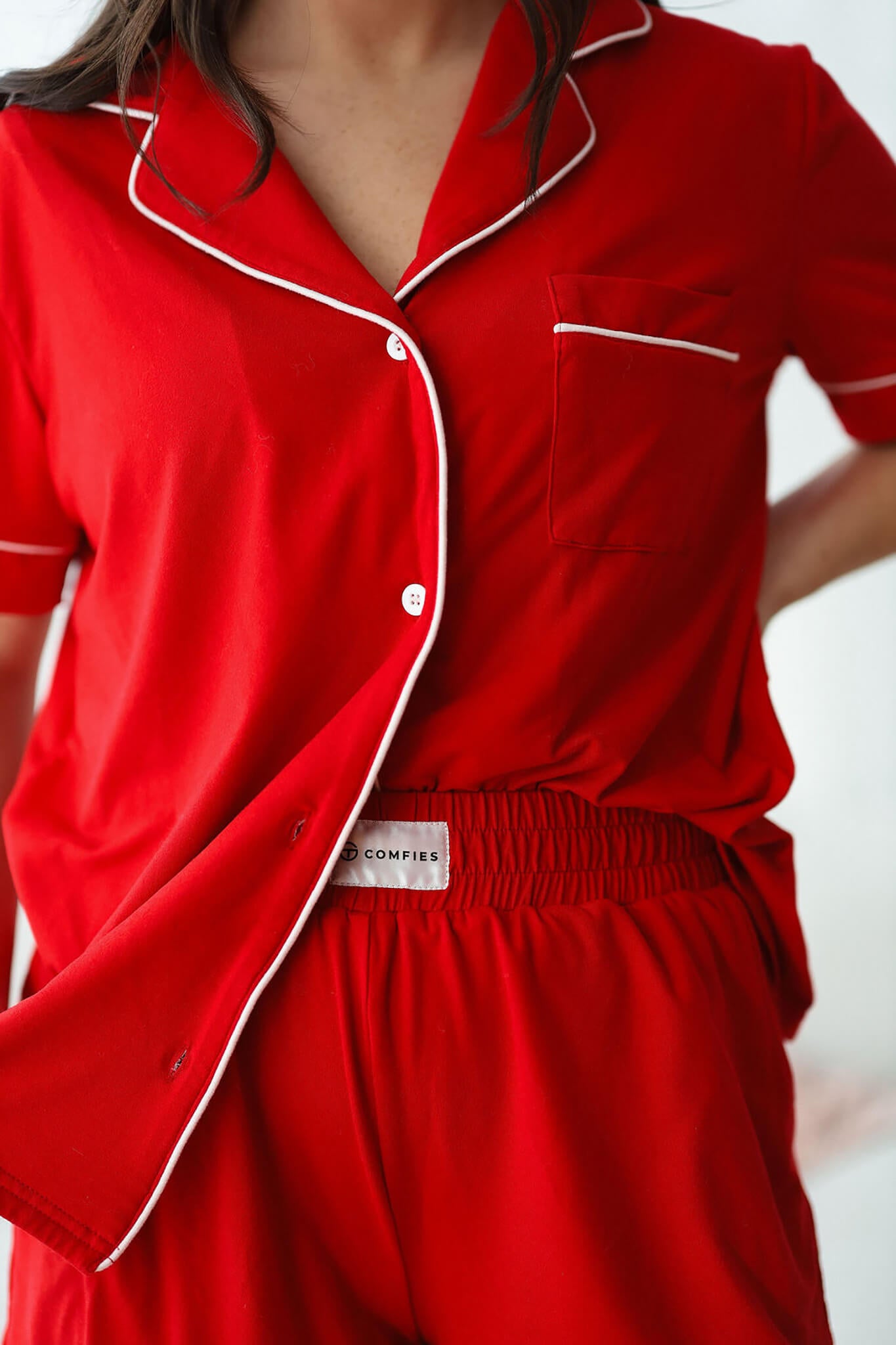 Model wearing red short sleeve pajama top and shorts with white piping, seated on a sofa in a relaxed setting.